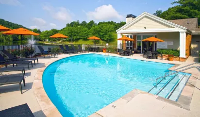 Sunny outdoor swimming pool with lounge chairs, orange umbrellas, a clubhouse, and a lake with a fountain in the background.