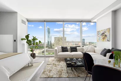 Bright modern studio apartment with floor-to-ceiling windows offering a city skyline view. The space features a white bed on the left, a large cream sectional sofa with black and gray pillows, a patterned area rug, and a black coffee table with magazines and decor. A dining table with black chairs and a vase of flowers is on the right, and a tall green plant adds a natural touch near the window. The walls are white with a colorful abstract painting, and the room is filled with natural light.