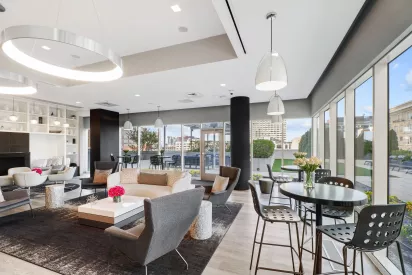 Modern, light-filled residential lounge with contemporary furniture, including sofas, armchairs, and café-style tables, large floor-to-ceiling windows, pendant lighting, and views of an outdoor terrace and city buildings.