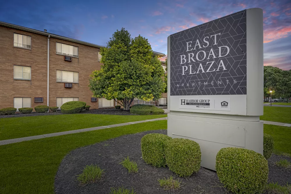 Graphic sign for East Broad Plaza Apartments in front of a brick building with a green lawn under a sunset sky.