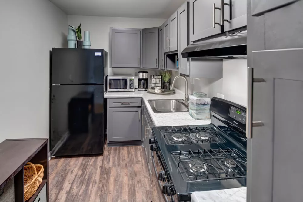 Modern L-shaped kitchen with grey cabinets, white countertops, black refrigerator, microwave, and gas stove.