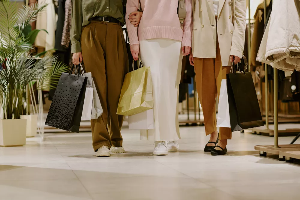 Three shoppers, visible from the waist down, walk arm-in-arm in a clothing store with shopping bags.