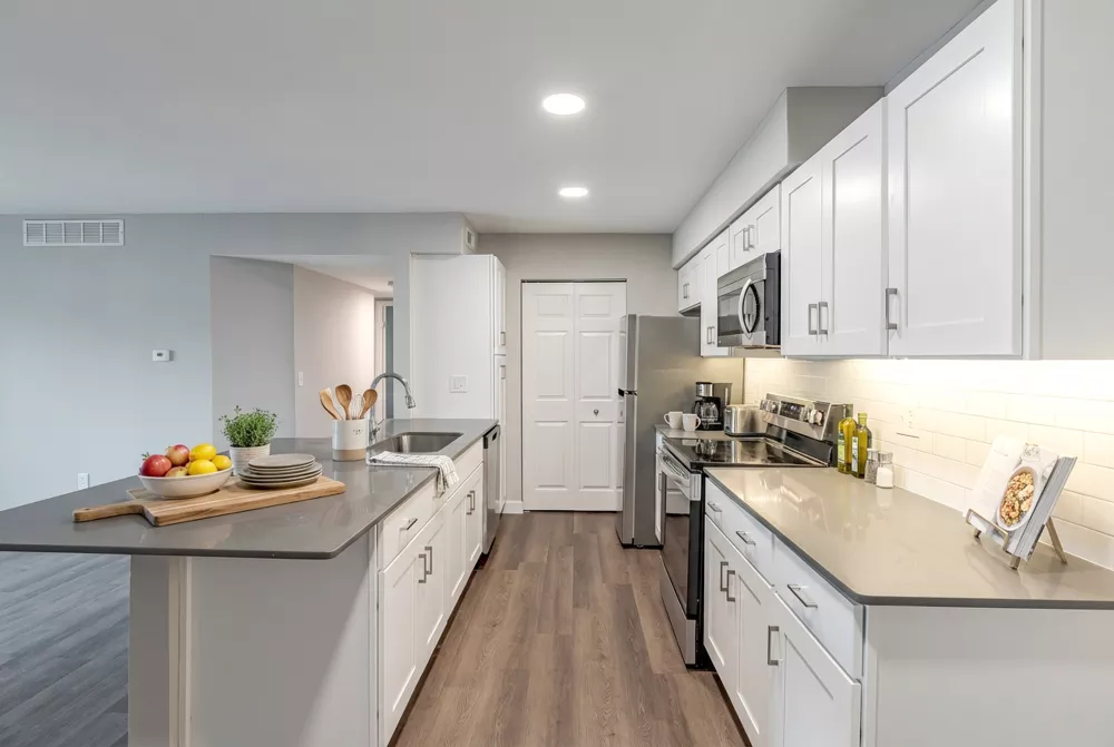 Modern kitchen with white cabinets, grey countertops, stainless steel appliances, and an island featuring a sink and fruit bowl.