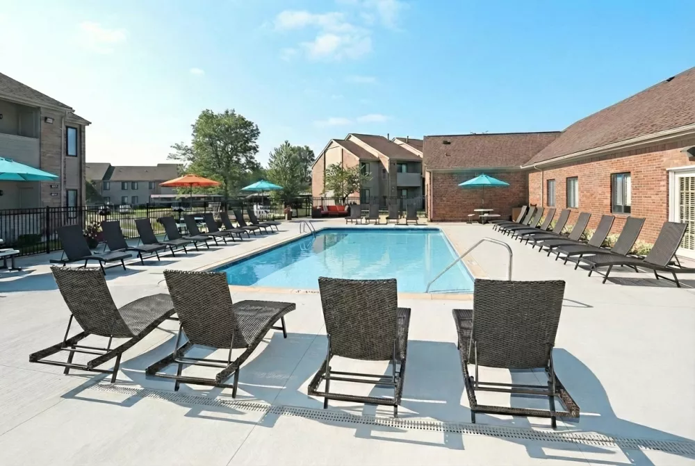 Sunny outdoor swimming pool with numerous lounge chairs, surrounded by apartment buildings under a clear blue sky.