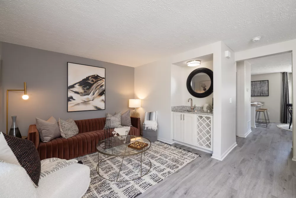 Modern living room with a burnt orange sofa, abstract art, coffee table, rug, and a built-in wet bar with a round mirror.