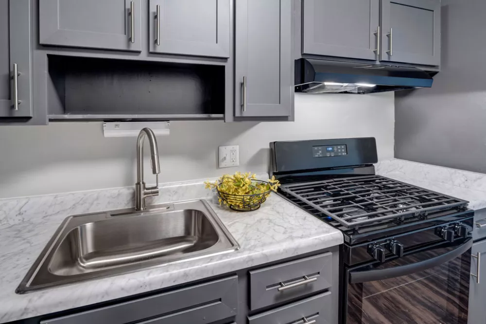 Kitchen with gray cabinets, white counter, stainless steel sink, black gas stove, and yellow flowers.
