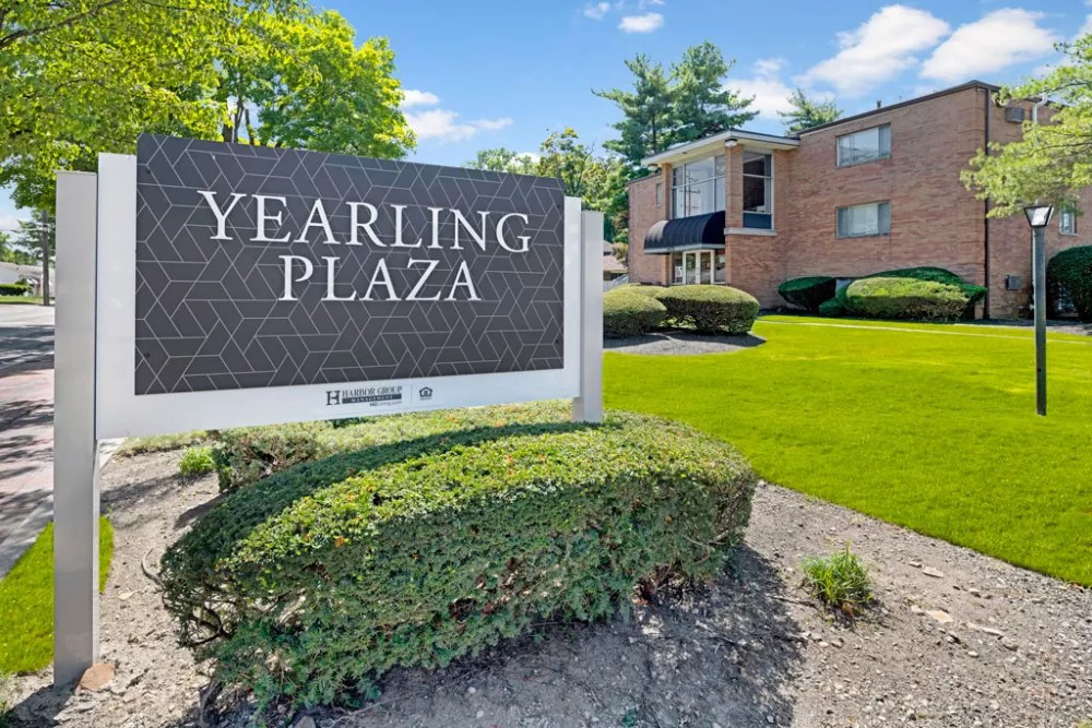 Sign for "YEARLING PLAZA" with geometric pattern, in front of a light-colored apartment building surrounded by green lawn.