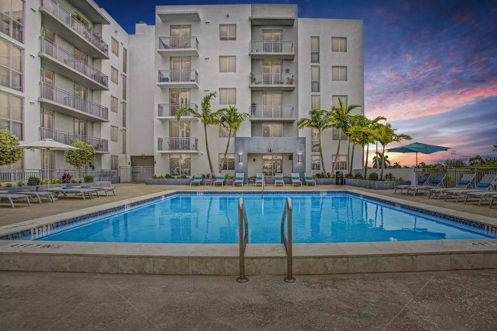Resort-style swimming pool with palm trees and lounge chairs in front of a modern white apartment building at sunset.