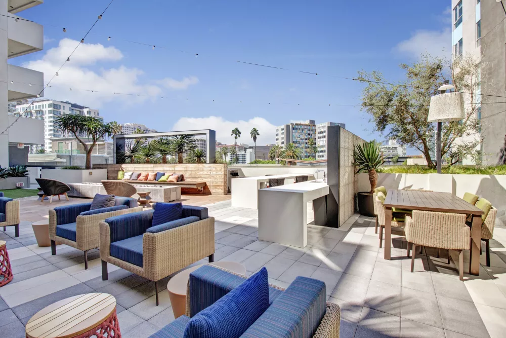 Rooftop patio with various seating areas, an outdoor dining table, and city buildings under a bright blue sky.