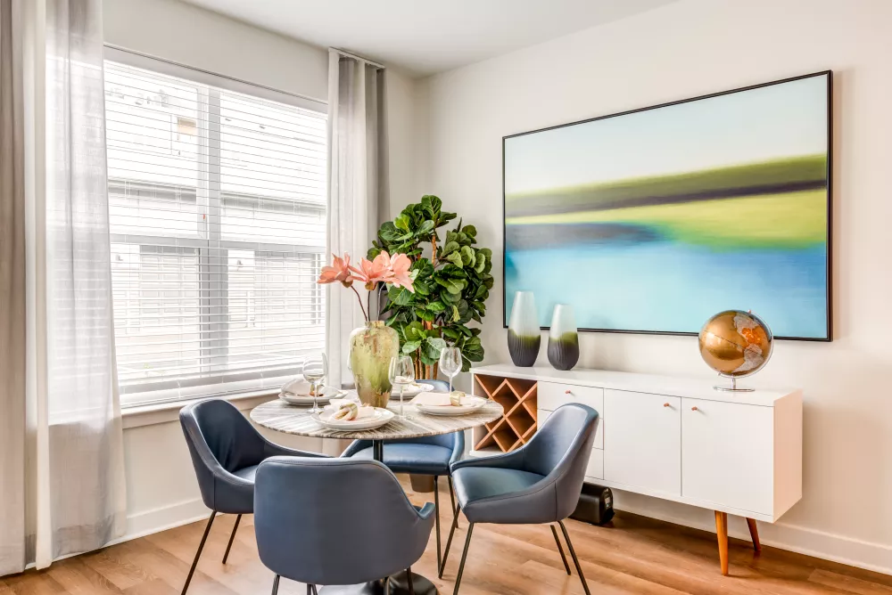 Modern dining area with a round marble table, blue chairs, a white credenza, abstract painting, and large window.