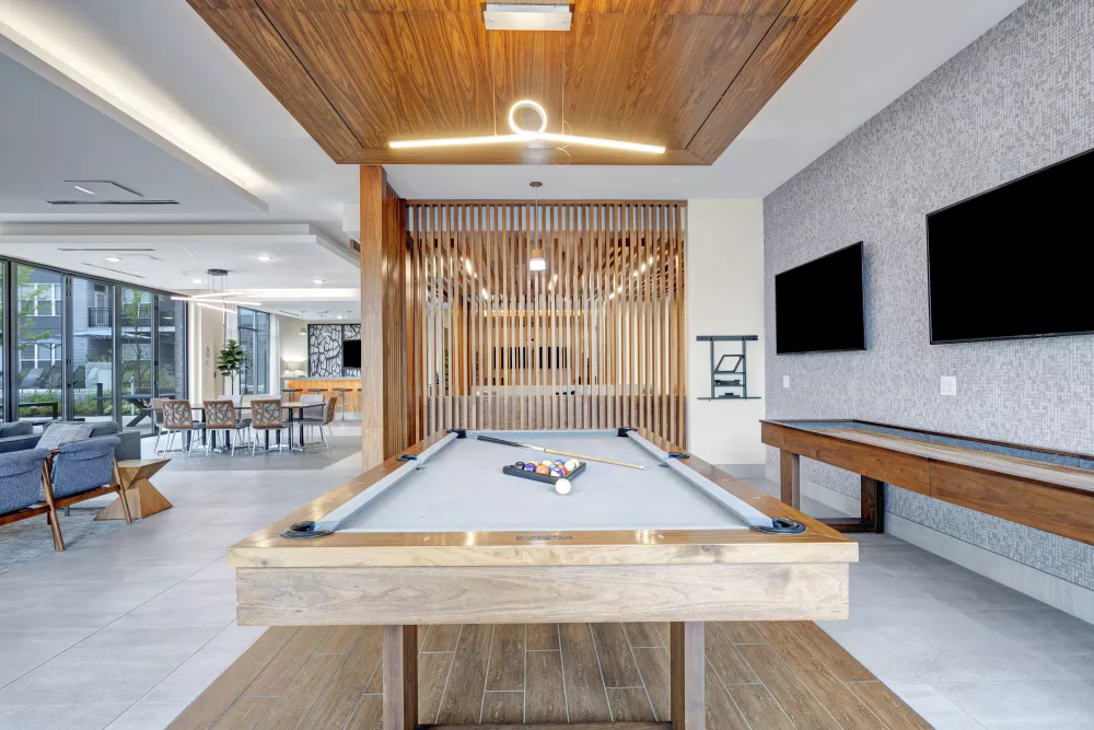 Modern lounge featuring a pool table with balls, a shuffleboard, and TVs. Wood slat wall and stylish ceiling lights. Seating area in background.