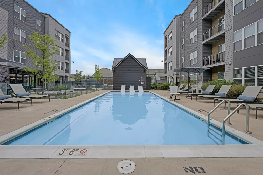 Outdoor swimming pool with lounge chairs between two modern apartment buildings and a dark gabled clubhouse under a blue sky.