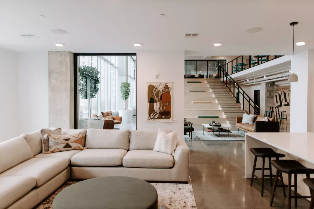 Modern open-plan living room with cream L-shaped sofa, round ottoman, kitchen island, and contemporary staircase.