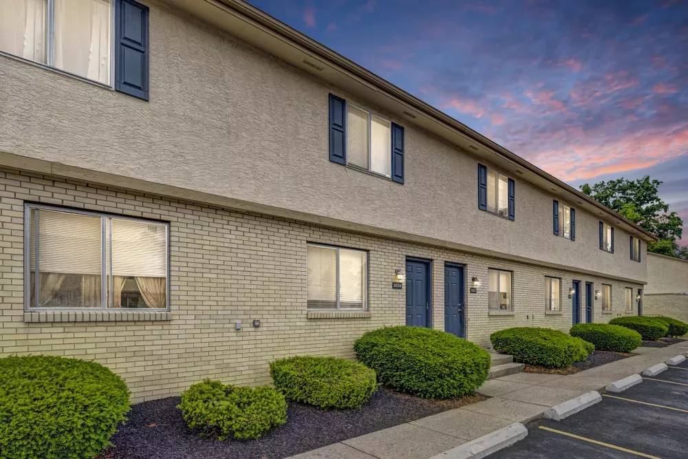 Two-story apartment building with tan facade, dark blue doors, and shutters. Green bushes line the front under a colorful sunset sky.