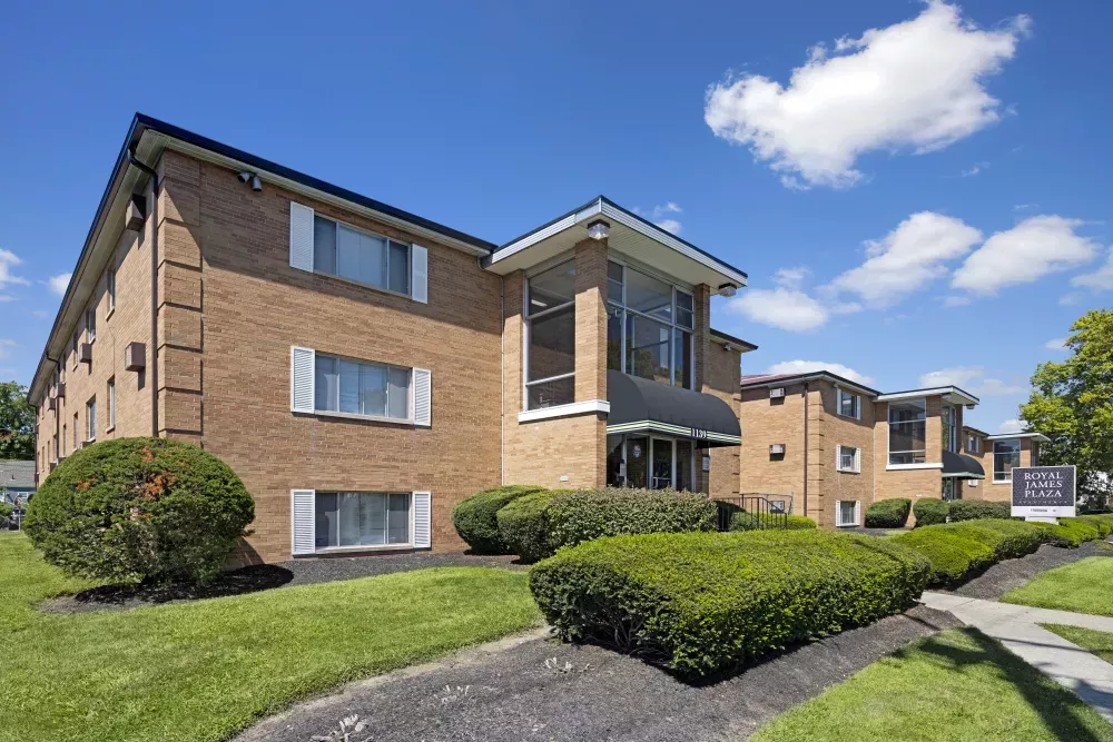 Brick apartment building with black awnings, green landscaping, and a clear blue sky.