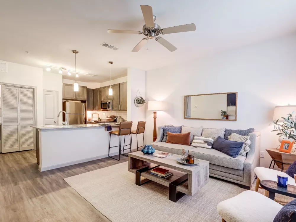 Bright open-concept living room with a gray sofa, wooden coffee table, and an adjacent kitchen island with bar stools.