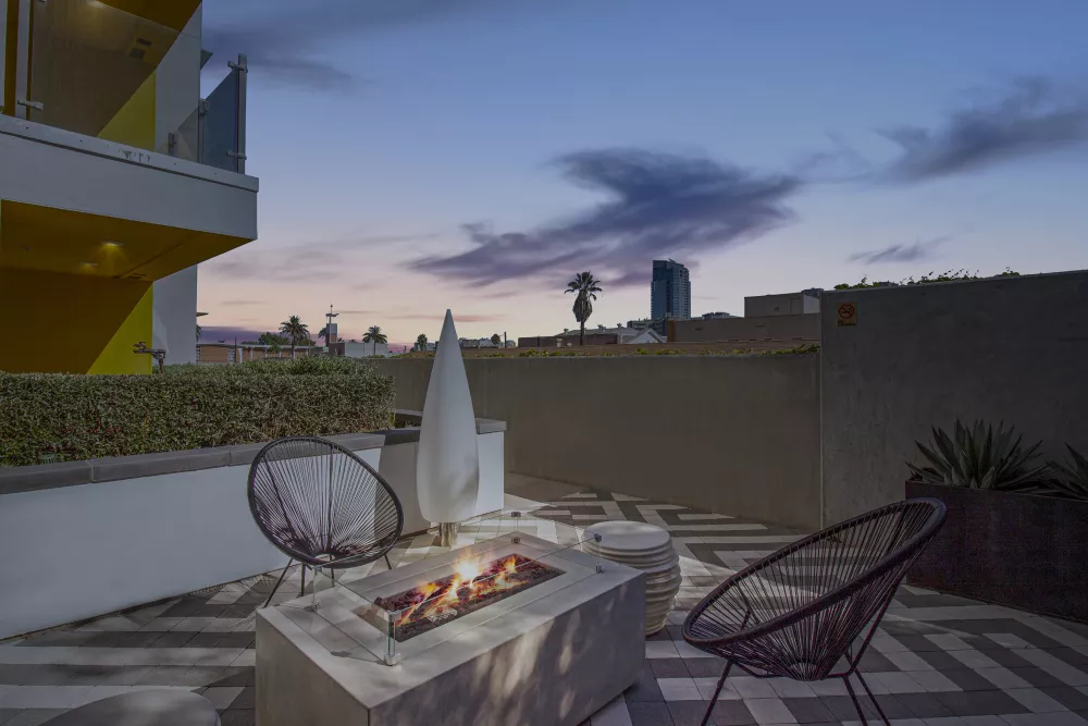 Dusk view of an outdoor patio with a lit fire pit, two chairs, and a city skyline under a twilight sky.