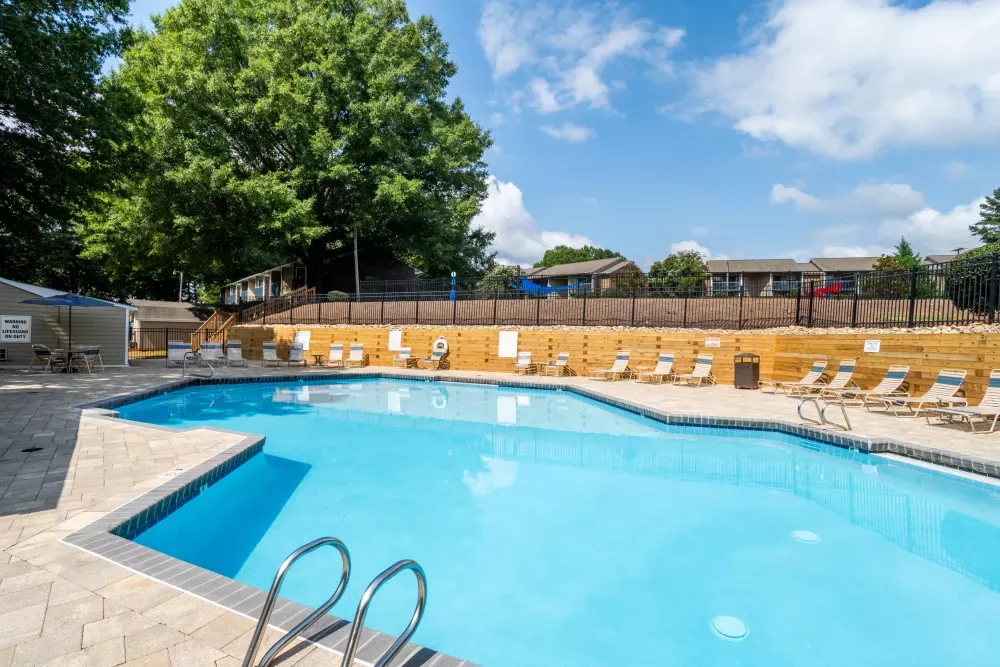 Sunny outdoor swimming pool with lounge chairs, a wooden fence, and apartment buildings under a blue sky.