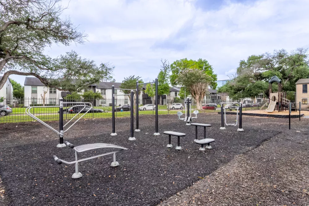 Outdoor fitness park with various exercise equipment on dark rubber surfacing, surrounded by trees and apartment buildings.