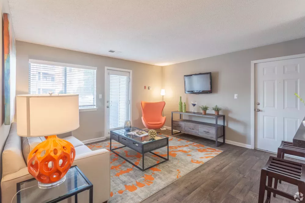 Modern living room with a light sofa, orange accent chair, coffee table on a gray and orange rug, and wall-mounted TV.