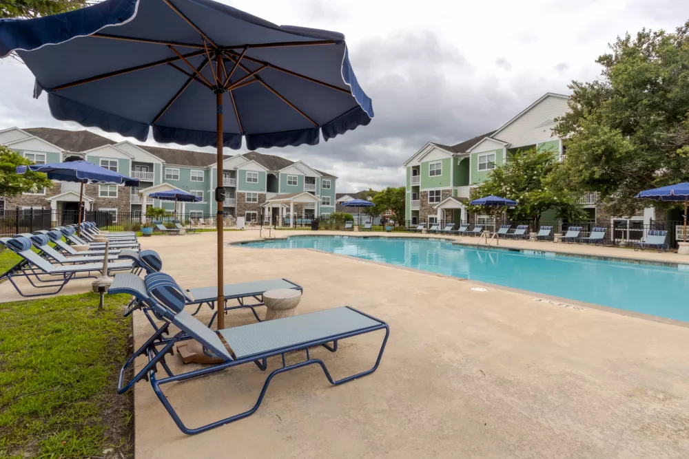 Residential swimming pool with blue lounge chairs, umbrellas, and apartment buildings on a cloudy day.