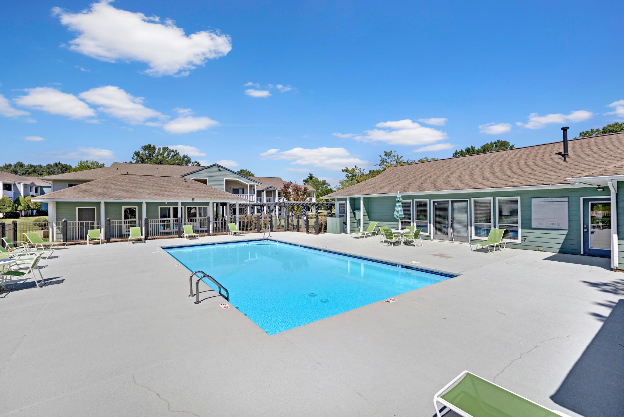 An outdoor swimming pool surrounded by green lounge chairs and a light blue clubhouse under a bright blue sky with fluffy white clouds.