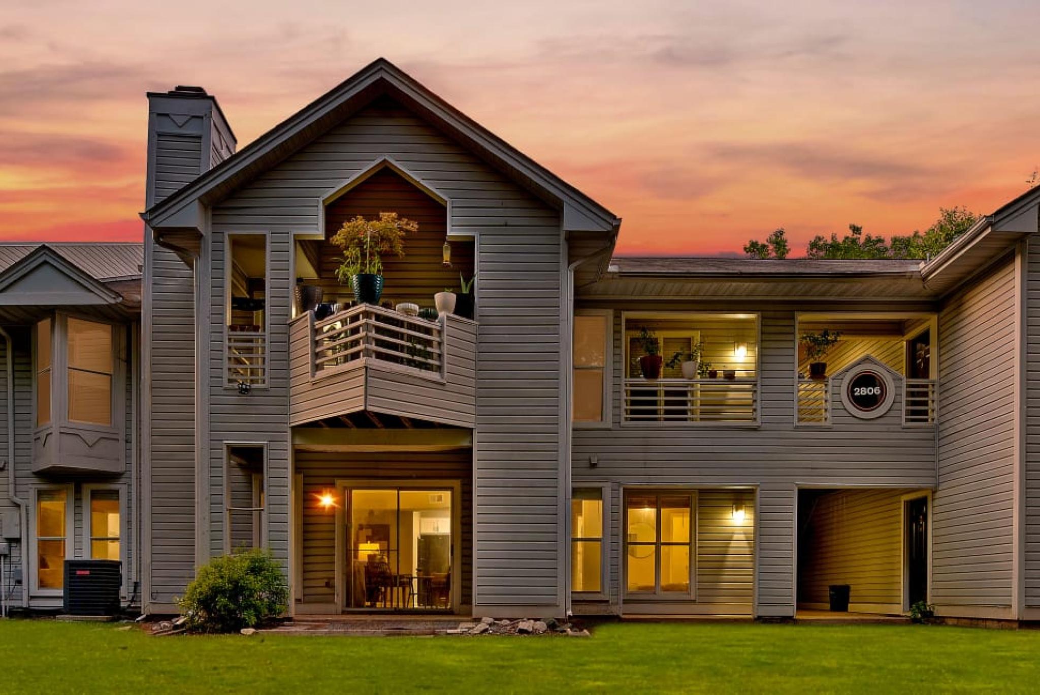 A two-story residential building with balconies, surrounded by greenery, set against a vibrant sunset sky.