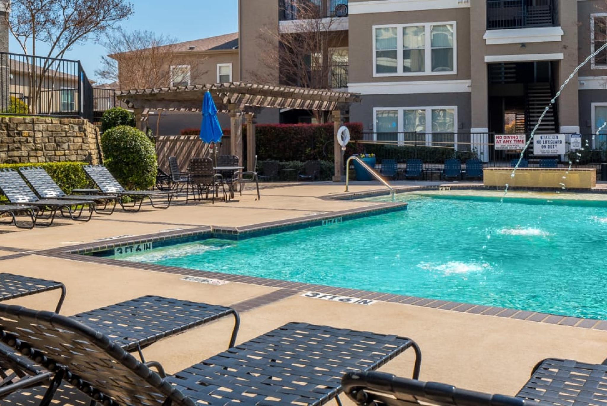 Outdoor swimming pool area with lounge chairs, fountains, and a shaded seating space.