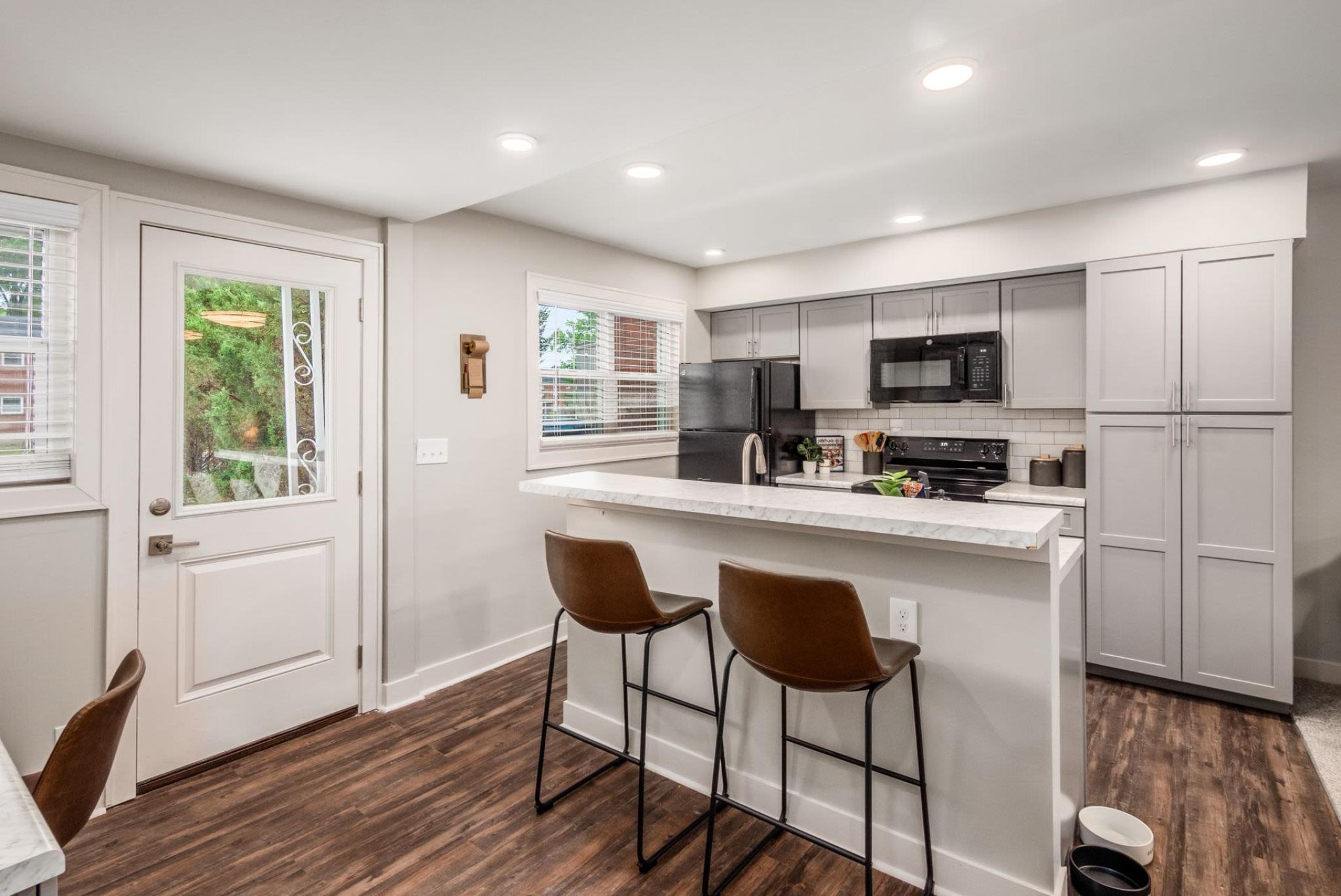 A modern kitchen with gray cabinetry, a marble countertop breakfast bar, and bar stools, complemented by hardwood floors and ample natural light from large windows.