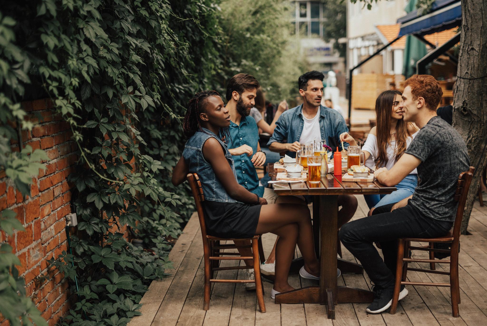 A group of friends enjoys food and drinks together at an outdoor restaurant patio surrounded by greenery.