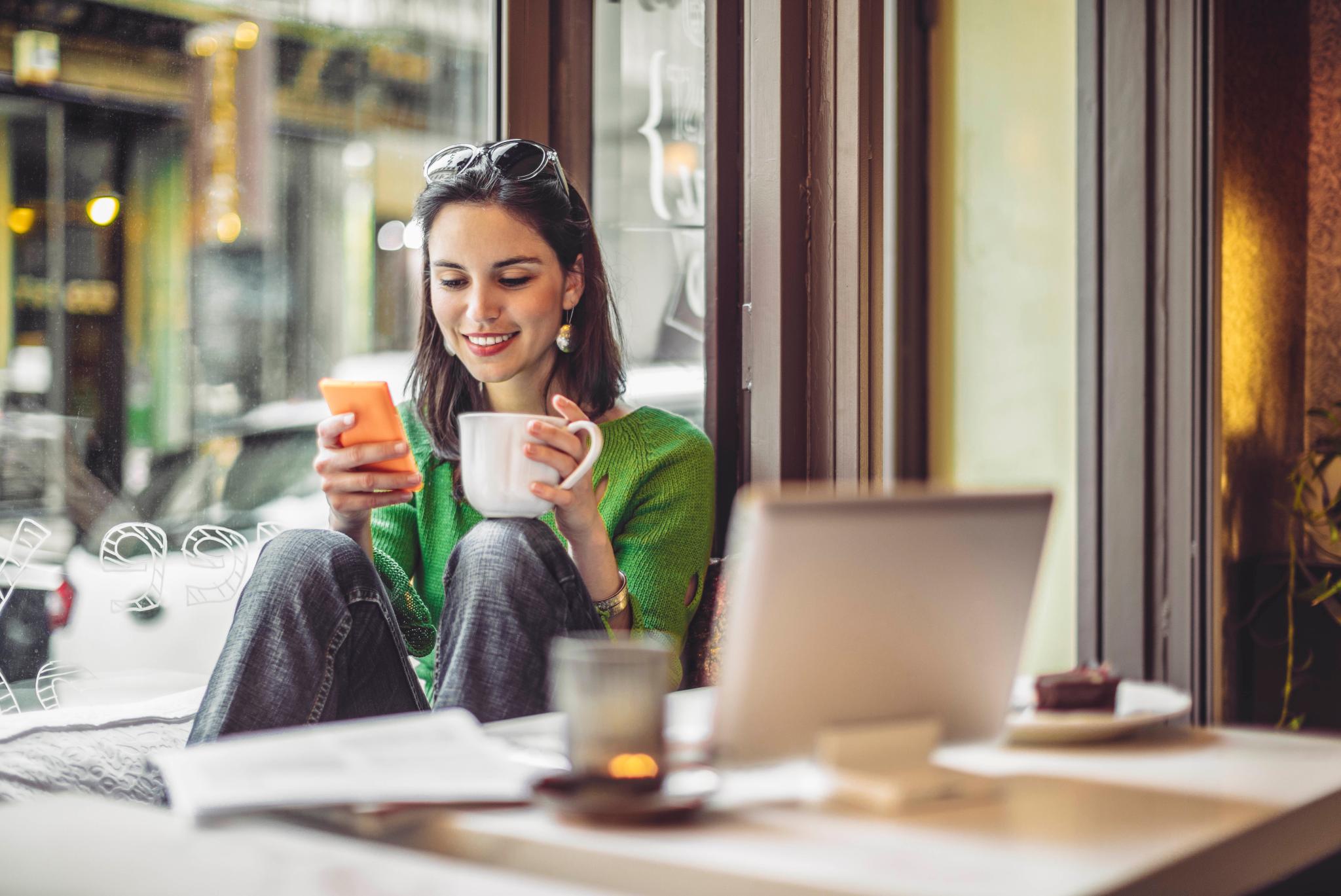 A woman sitting by a cafe window, smiling as she looks at her phone while holding a coffee mug in a cozy and relaxed setting.