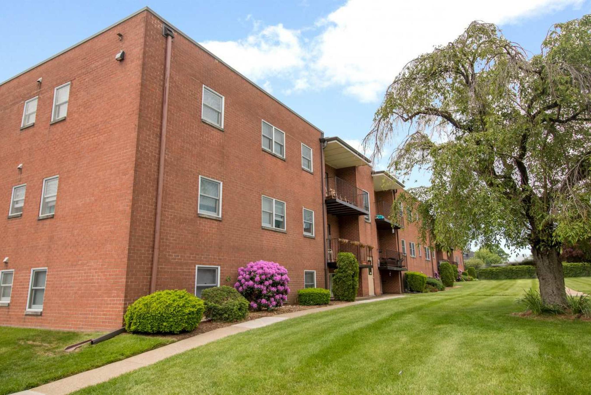 A red brick apartment building with well-maintained green lawns, colorful bushes, and a large tree in the foreground under a partly cloudy sky.