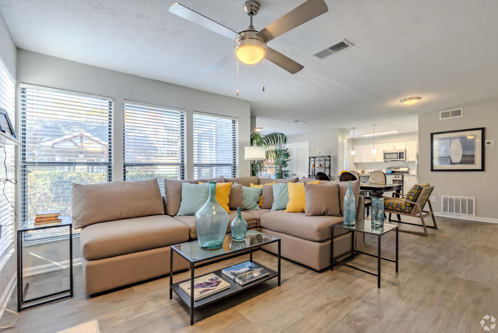 A spacious living room featuring a large sectional sofa with colorful throw pillows, a ceiling fan, and large windows letting in ample natural light.