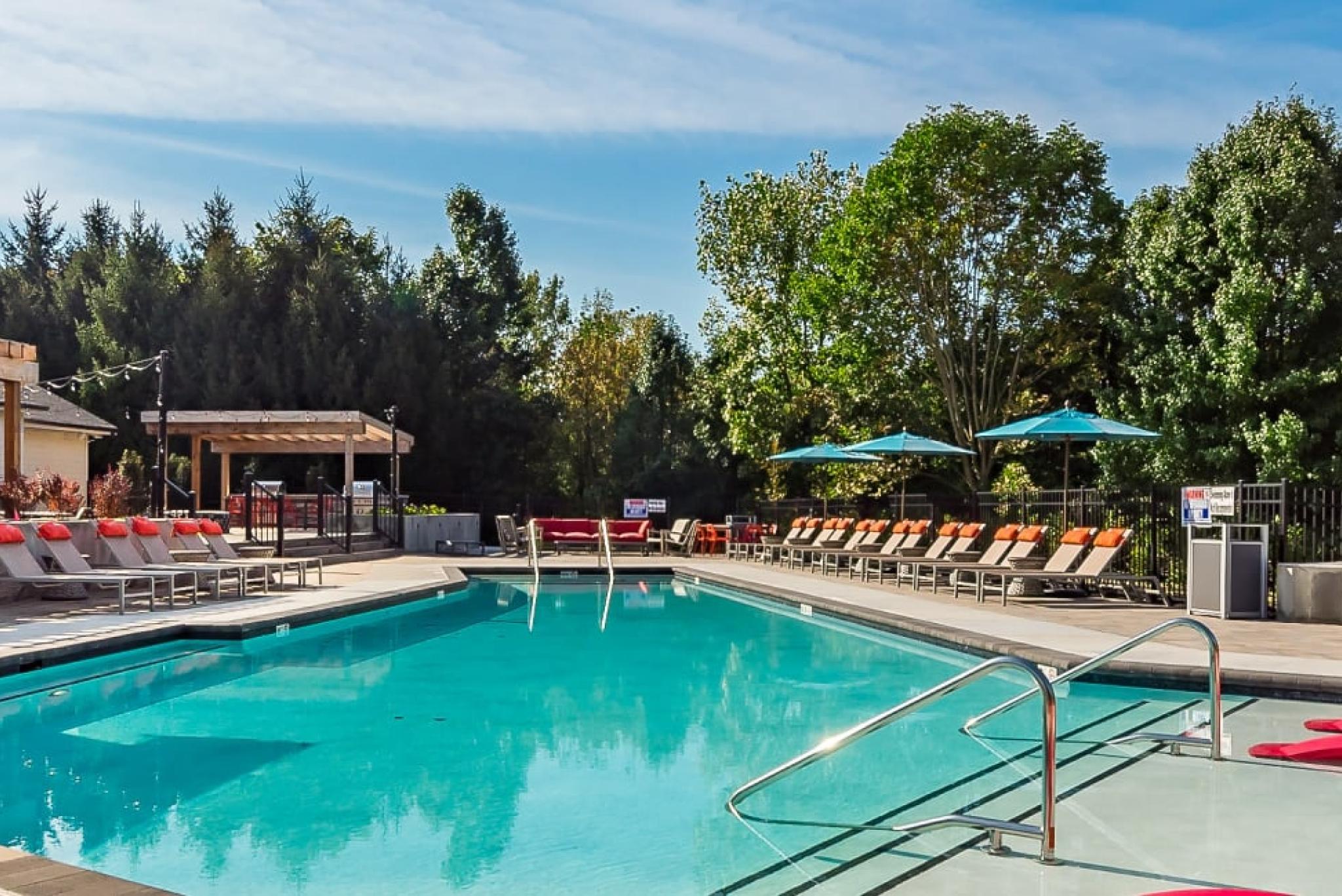 A sparkling outdoor swimming pool surrounded by lounge chairs, umbrellas, and lush greenery, offering a relaxing atmosphere at The Garratt Apartments.
