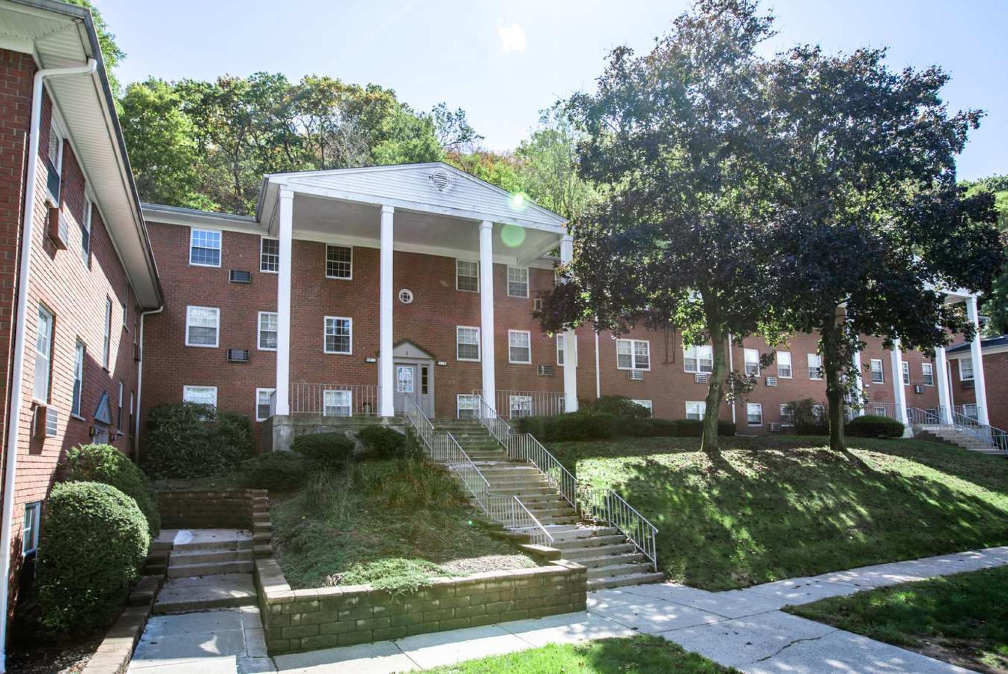 A charming brick apartment building at The Manors Apartments, surrounded by lush greenery and featuring classic white columns at the entrance, offering a welcoming and serene living environment.