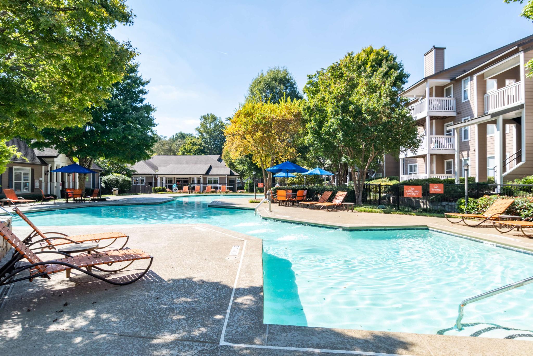 Resort-style swimming pool surrounded by orange lounge chairs and shaded seating under blue umbrellas.