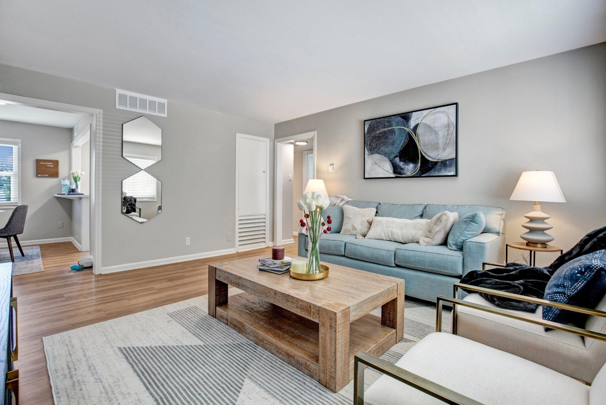 A stylish and cozy living room at Alcove at Seahurst, featuring a soft blue sofa adorned with decorative pillows, a rustic wooden coffee table with a floral centerpiece, and two modern accent chairs with gold metal frames. The space is illuminated by warm table lamps, and abstract wall art enhances the contemporary decor. An open doorway leads to a small dining area, and large windows bring in natural light, creating an inviting atmosphere. ​​