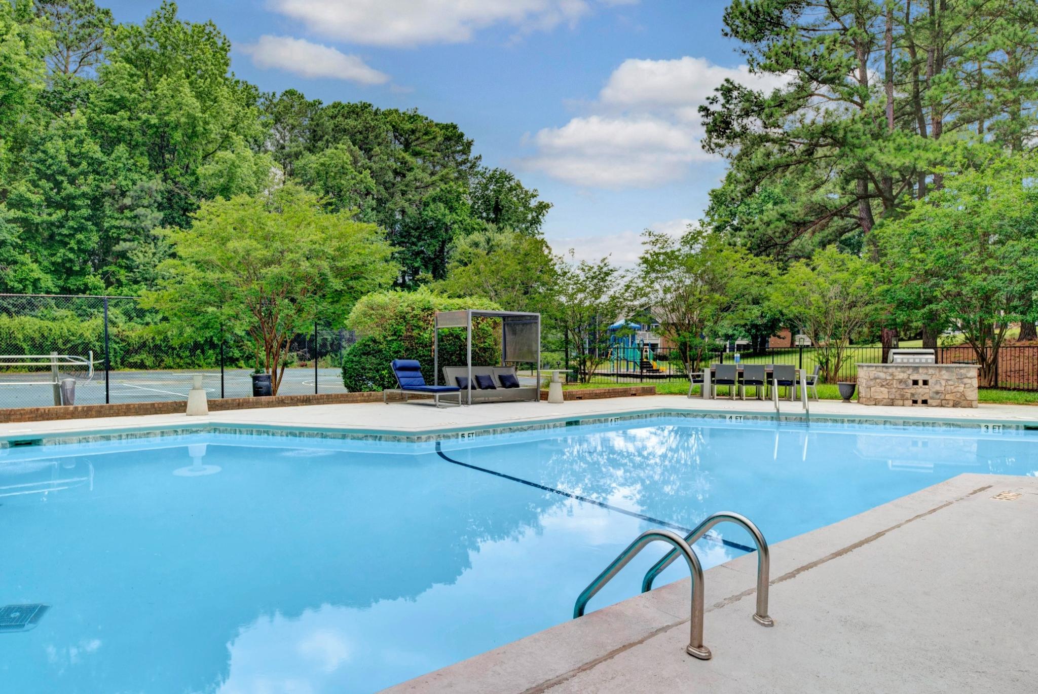 A serene outdoor pool area at Aria North Hills, surrounded by lush greenery and tall trees. The pool features stainless steel handrails, lounge chairs, and a shaded cabana area for relaxation. A tennis court and a children's playground can be seen in the background, offering a variety of recreational amenities. 