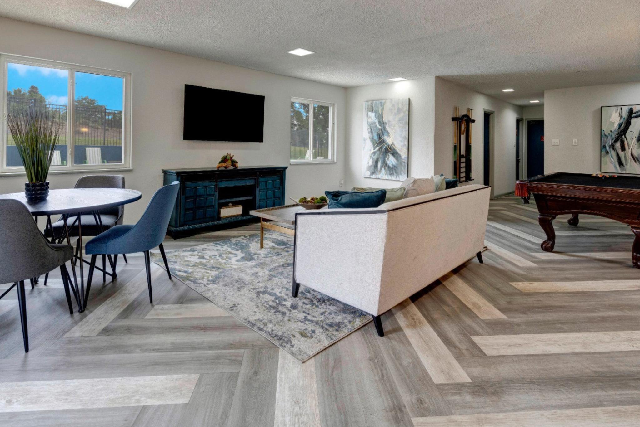 A stylish community lounge with a modern design, featuring a light beige sofa with blue accent pillows, a dark wood pool table with orange tassels, and a round dining table with blue chairs. The room includes a sleek entertainment center with a mounted flat-screen TV, large windows allowing natural light, and abstract art pieces on the walls. The flooring is arranged in a chevron pattern with shades of gray and beige. ​​