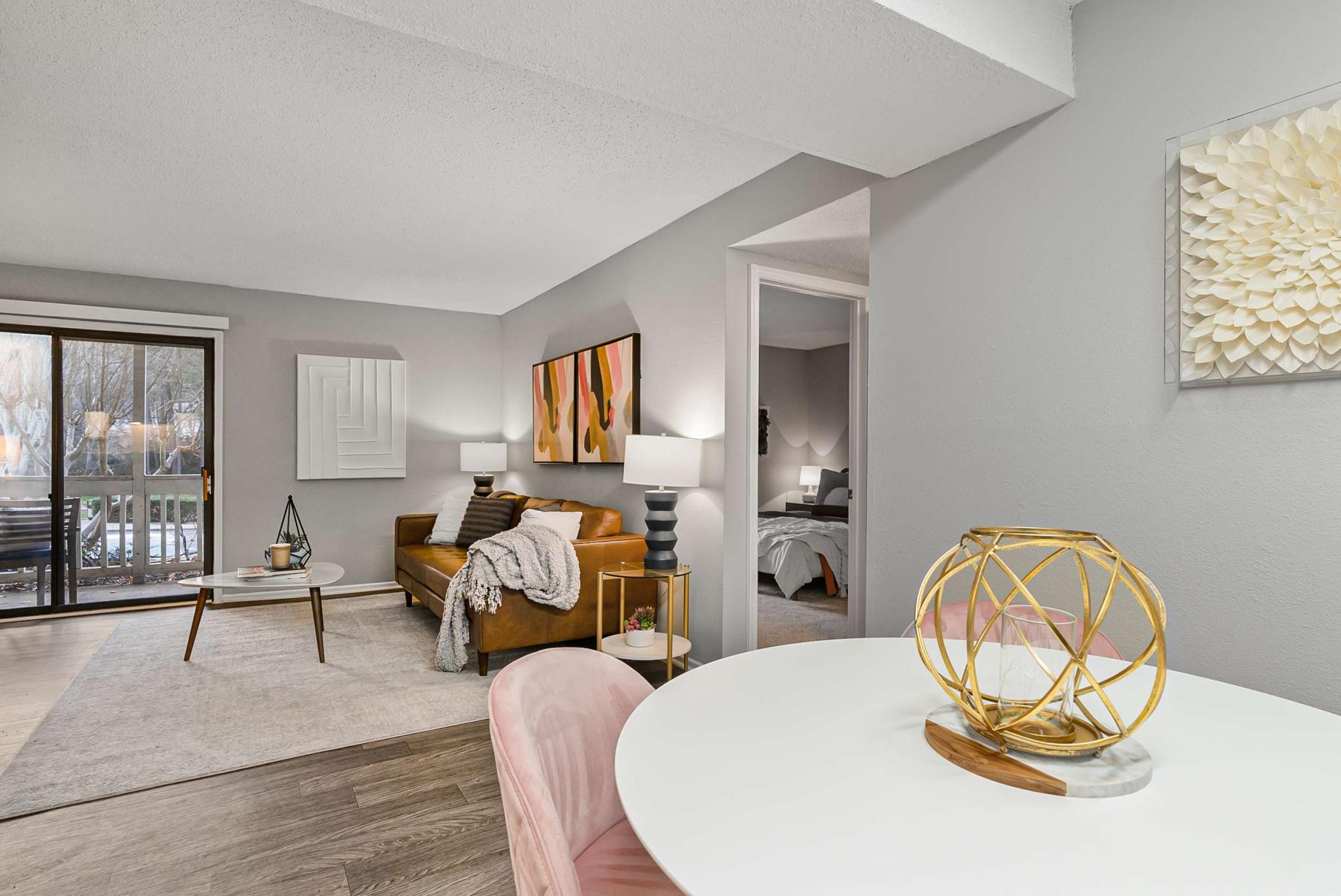 Modern apartment interior with a cozy leather sofa, abstract wall art, and a view of the bedroom and outdoor patio through sliding glass doors, accented by soft lighting and stylish decor.