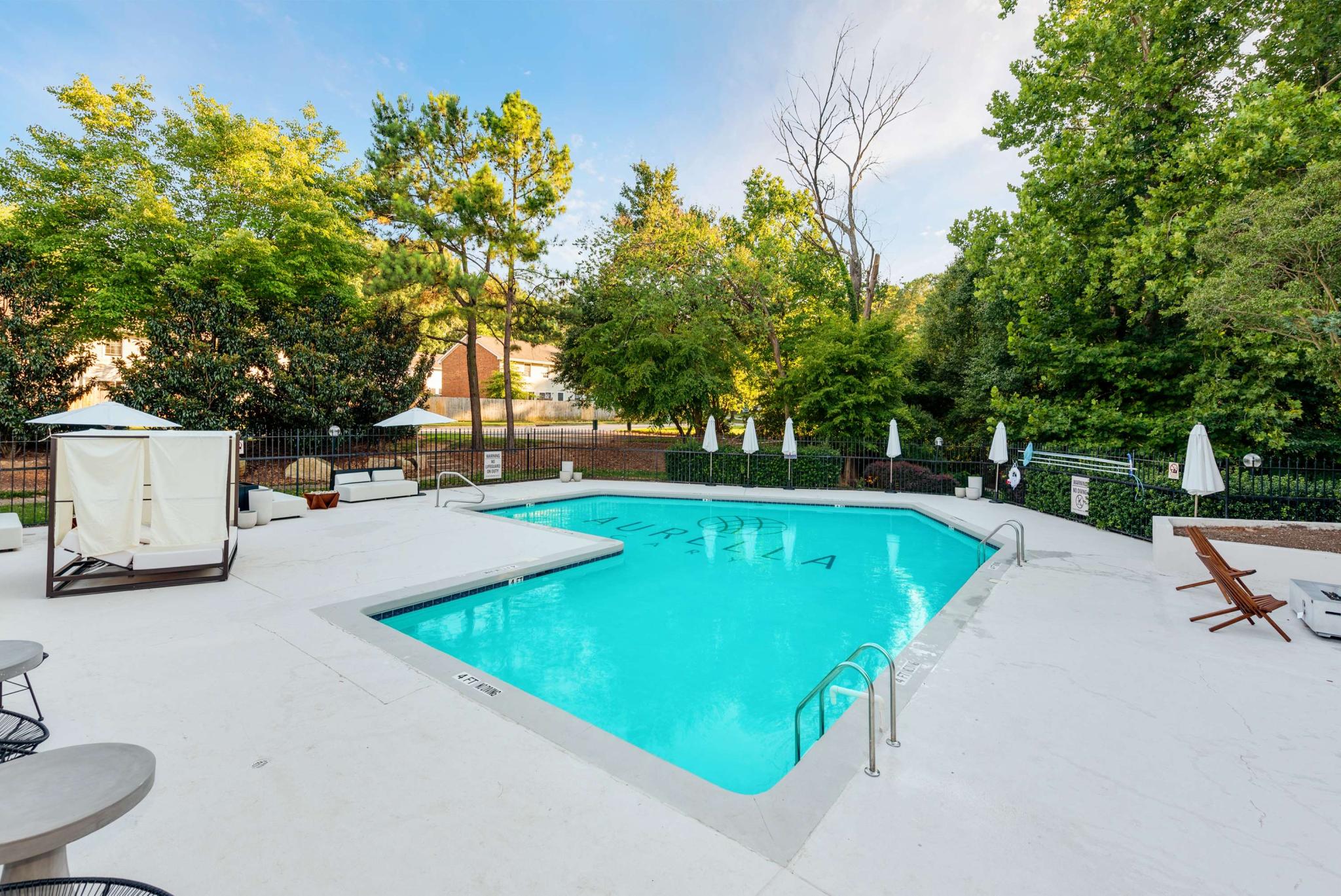 Resort-style swimming pool area featuring a bright turquoise pool, white poolside loungers and umbrellas, surrounded by lush greenery and a serene outdoor setting.