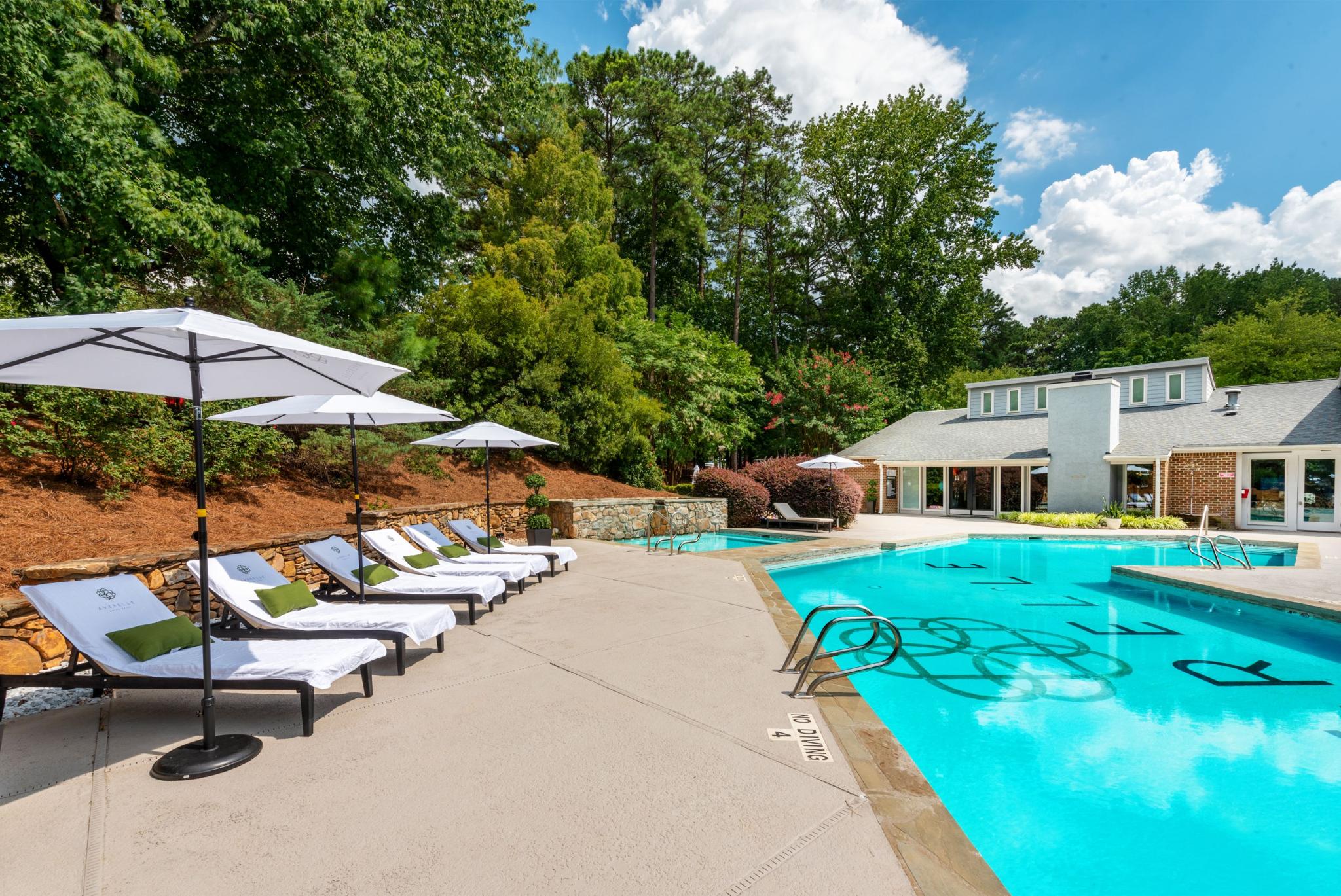 Inviting outdoor pool area with cushioned lounge chairs and white umbrellas, surrounded by lush landscaping and a clubhouse with large windows, offering a tranquil and stylish retreat.