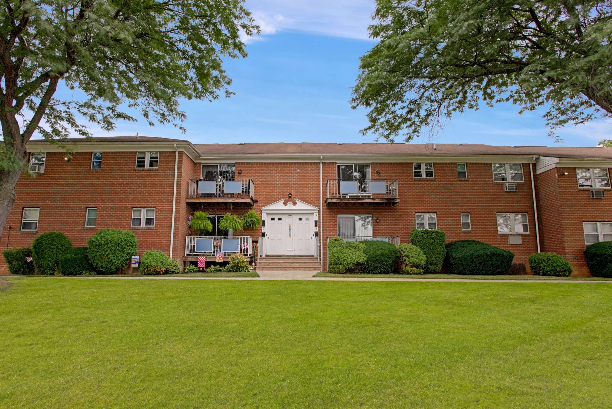 Charming brick apartment building with balconies and well-manicured landscaping, set against a backdrop of mature trees and a wide, green lawn under a clear blue sky.
