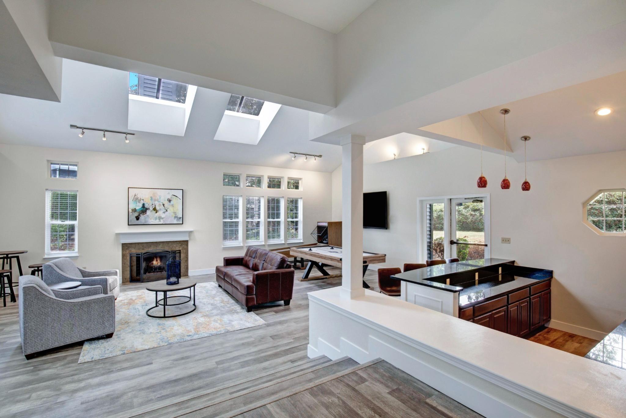 A spacious and modern clubhouse at Chandlers Bay featuring an open-concept design with high ceilings and skylights. The room is furnished with a brown leather sofa, gray armchairs, and a contemporary fireplace with a white mantel. A stylish wooden pool table is positioned near a wall-mounted TV, and a built-in bar area with granite countertops and red pendant lights adds to the upscale ambiance. Large windows and glass doors allow natural light to fill the space, offering views of the outdoor greenery. 