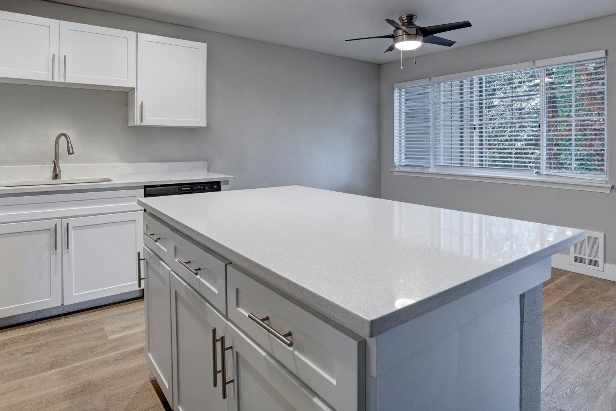 A modern kitchen at Chandlers Bay featuring sleek white cabinetry with silver hardware, quartz countertops, and a spacious center island with ample storage. The kitchen is equipped with a stainless steel sink and a high-arc faucet. Large windows with blinds allow natural light to brighten the space, and a ceiling fan with a built-in light fixture adds functionality. The open floor plan connects seamlessly to the adjacent living area, creating a contemporary and inviting atmosphere. 