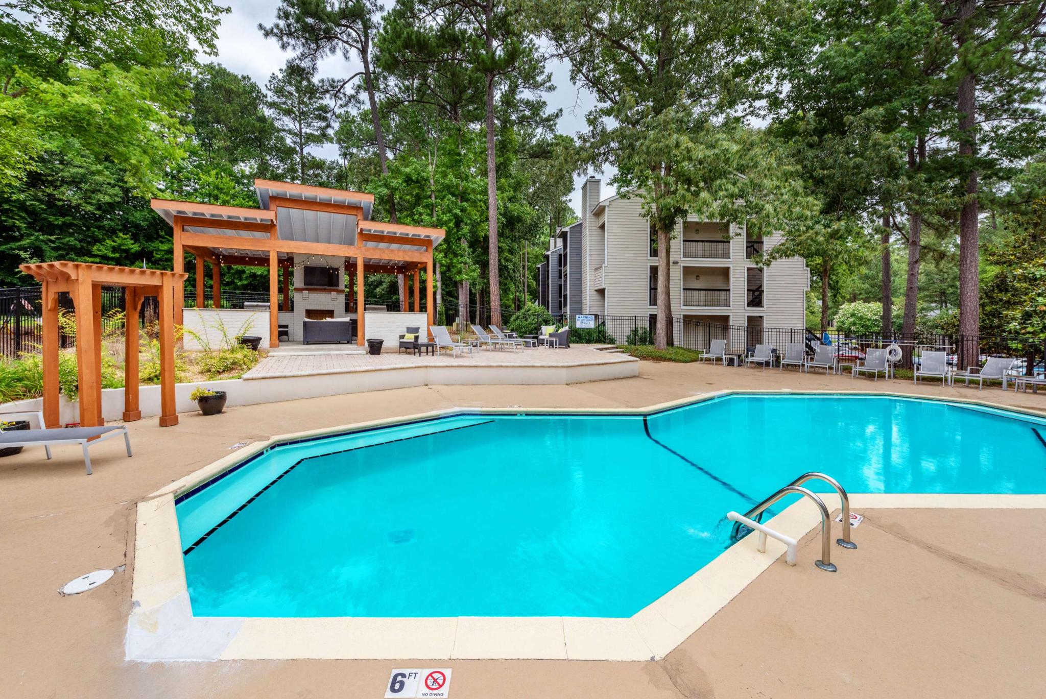 Hexagonal outdoor swimming pool surrounded by lounge chairs, with a modern wooden pergola and grill area set against a lush wooded backdrop and apartment buildings.