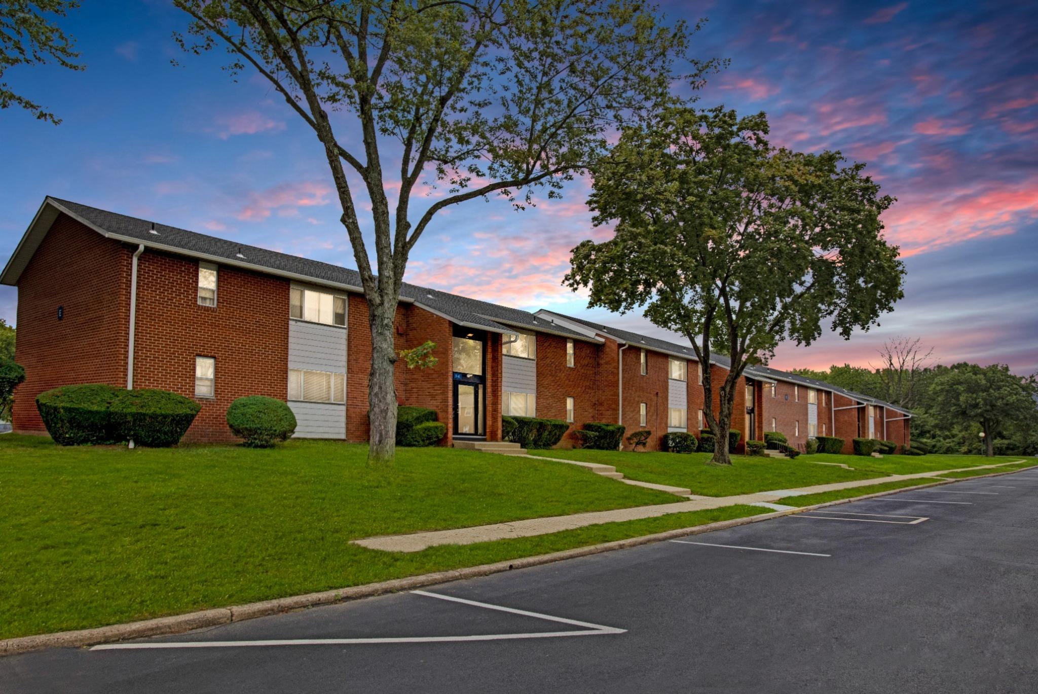 The exterior of Laurel Run Village Apartments showcases a well-maintained, two-story brick residential building with manicured green lawns and mature trees. The architecture features a mix of red brick and gray siding, with large windows and dark entrance doors. A spacious parking lot is in the foreground, and a sunset sky in the background adds a warm, inviting atmosphere. 
