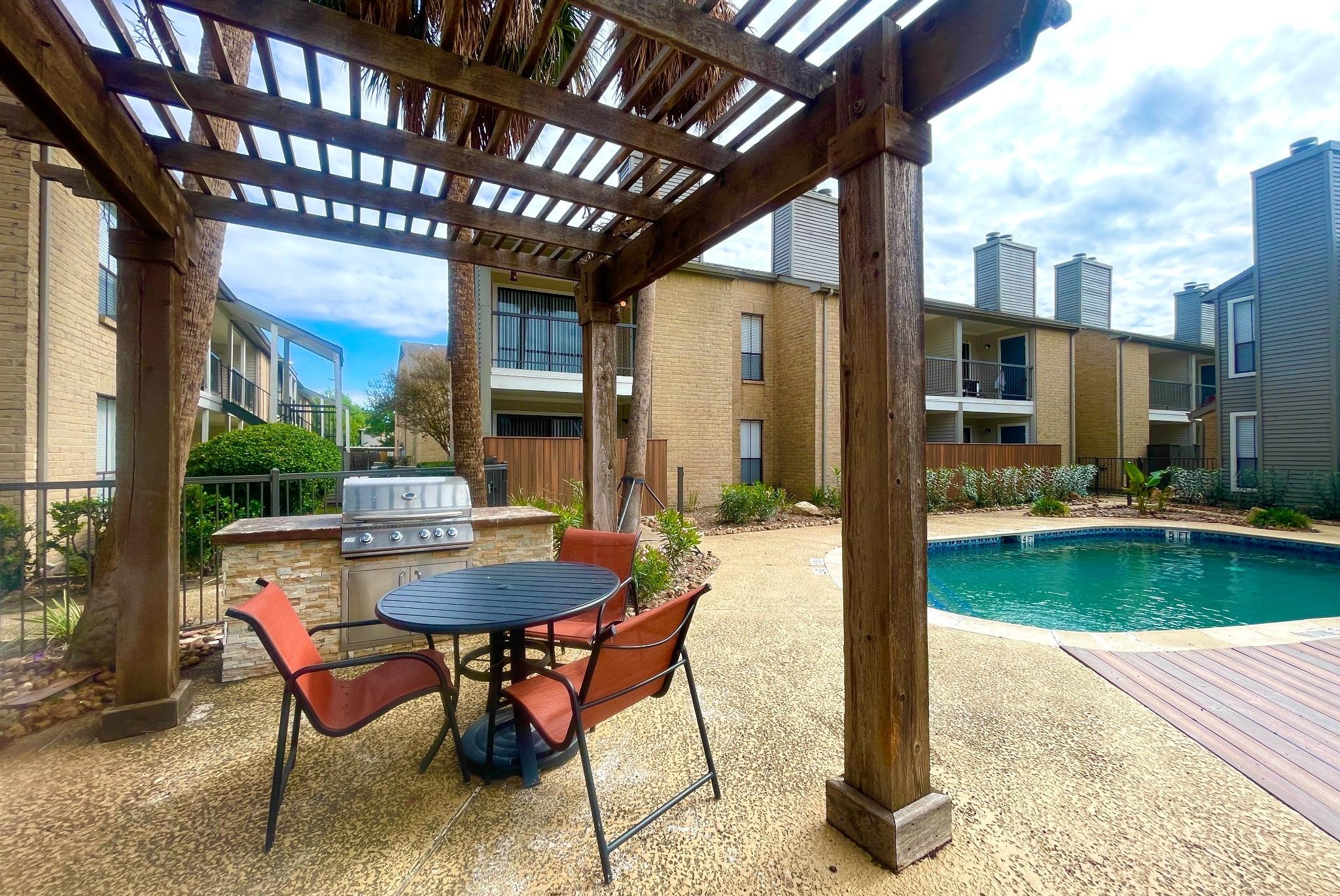 An inviting outdoor amenity space at Oaks of Westchase featuring a shaded pergola with a round dining table and four red chairs. Adjacent to the seating area is a built-in stainless steel grill for outdoor cooking. The area overlooks a sparkling swimming pool, surrounded by lush landscaping and modern apartment buildings with private balconies. The scene is framed by tall palm trees, creating a relaxed and resort-like atmosphere.