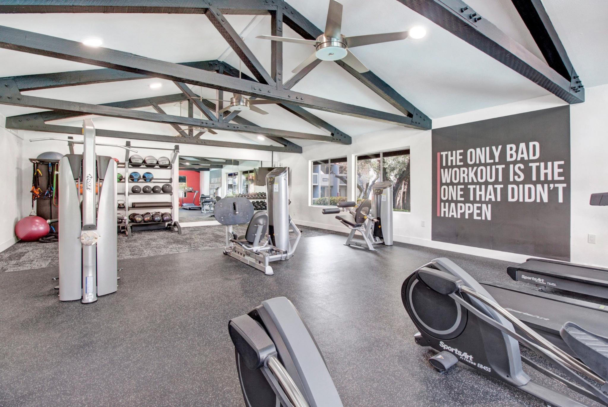 A modern fitness center at Olive Ridge Apartments, featuring high ceilings with exposed dark beams and large windows for natural light. The gym is equipped with state-of-the-art strength and cardio machines, free weights, and functional training accessories. A motivational wall mural reads, 'The only bad workout is the one that didn’t happen.'