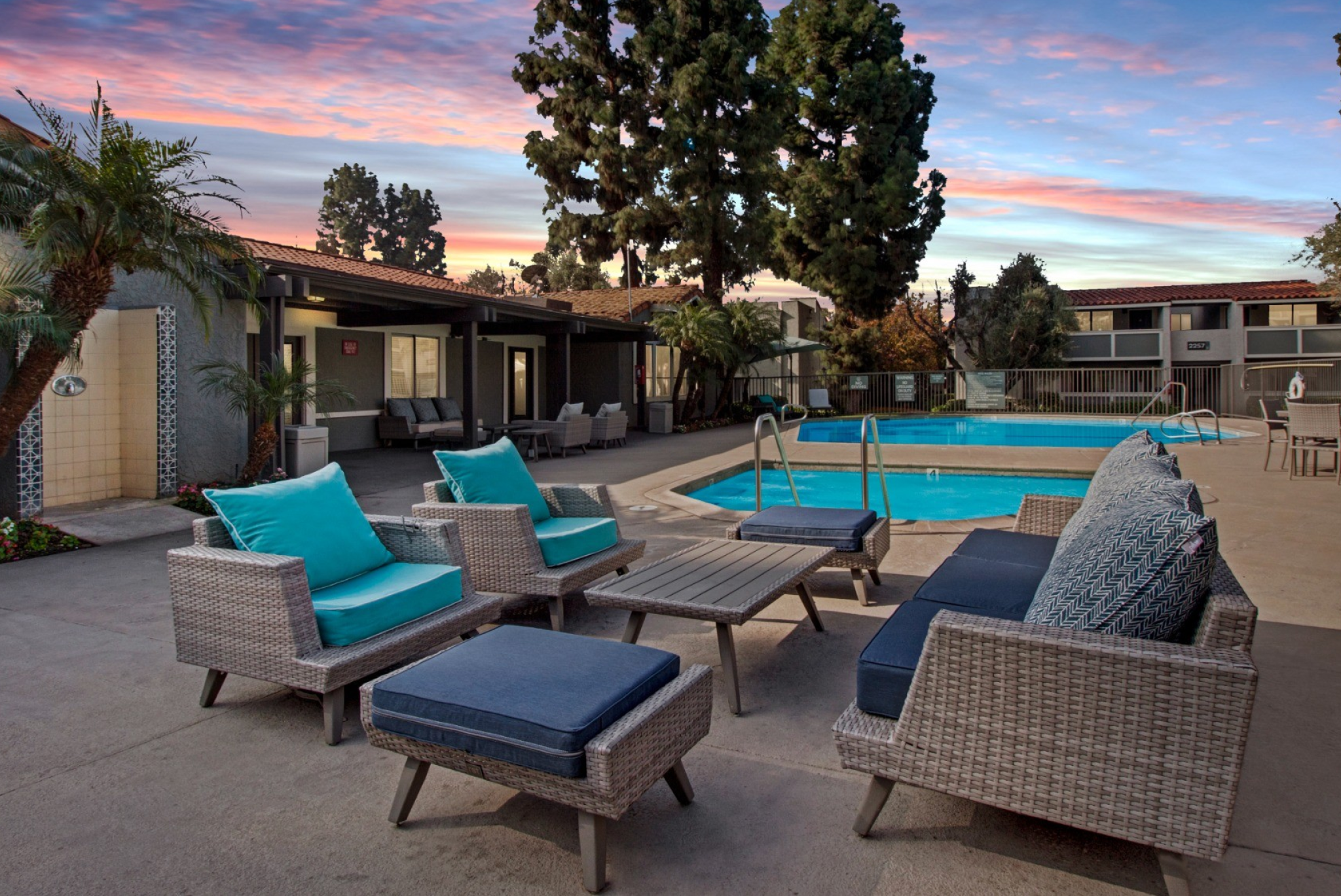 A resort-style swimming pool area at Olive Ridge Apartments captured during twilight. The space features woven wicker lounge chairs with plush blue and turquoise cushions, a communal seating area with ottomans, and a shimmering pool surrounded by lush landscaping. The evening sky is painted in shades of pink and blue, creating a serene atmosphere.